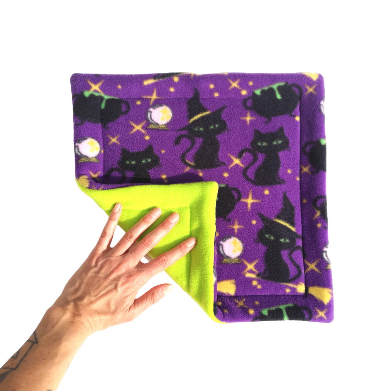 A hand holds a purple Halloween themed print fleece pet pee pad featuring cauldrons and black cats wearing witches hats with a like green reverse side