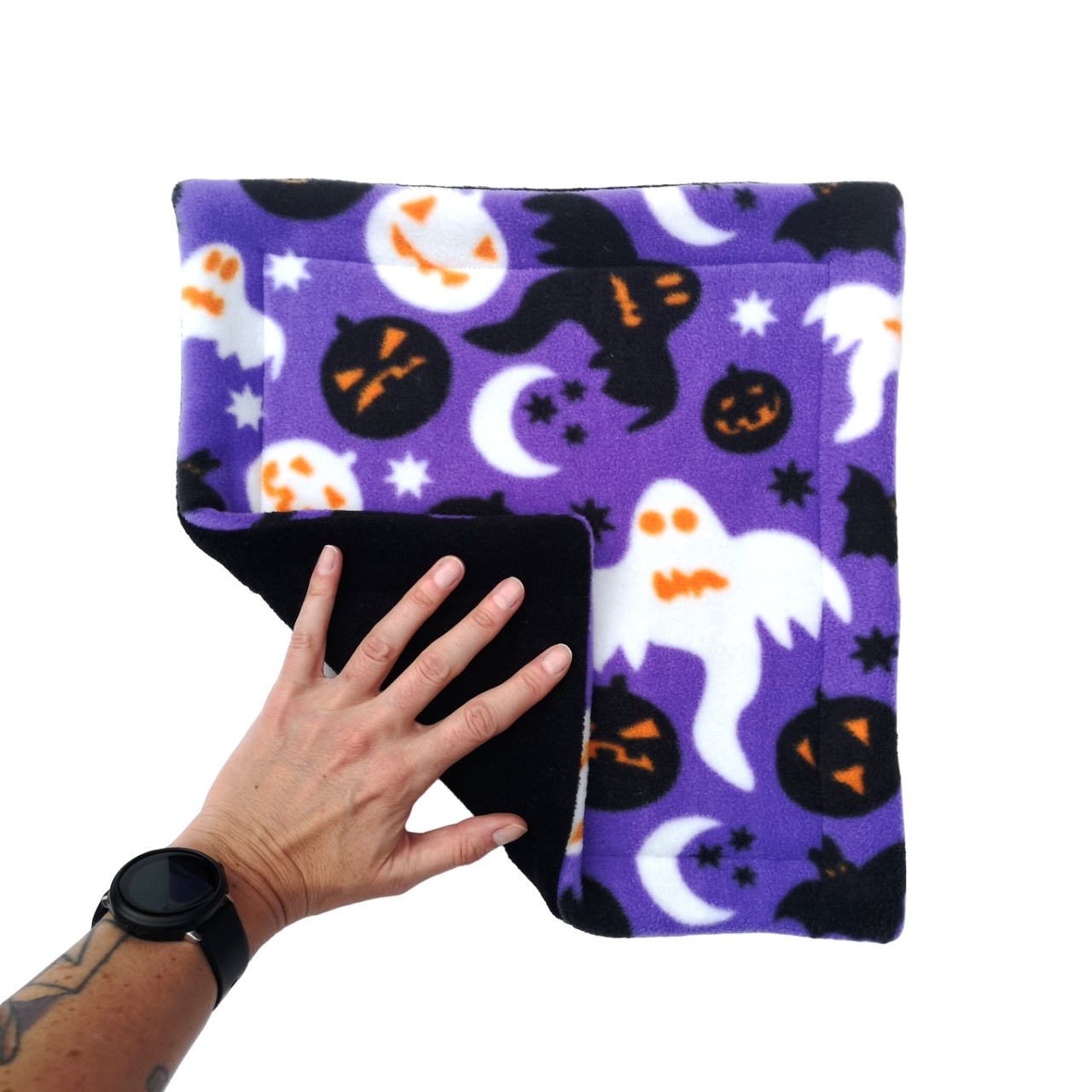 A hand holds a purple Halloween themed print fleece pet pee pad featuring ghosts, bats and pumpkins with a black reverse side