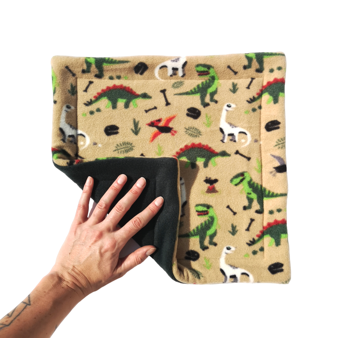 A hand holds a light brown dinosaur print fleece pet pee pad featuring green dinosaurs with a dark green reverse side