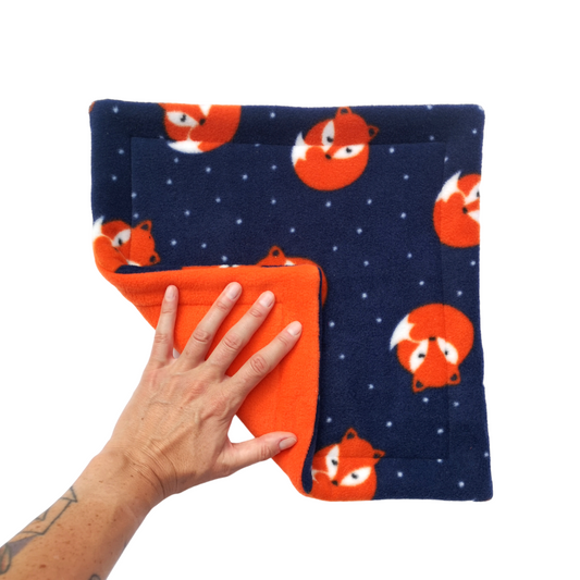A hand holds a dark blue fleece pet pee pad featuring orange sleeping foxes with an orange reverse side