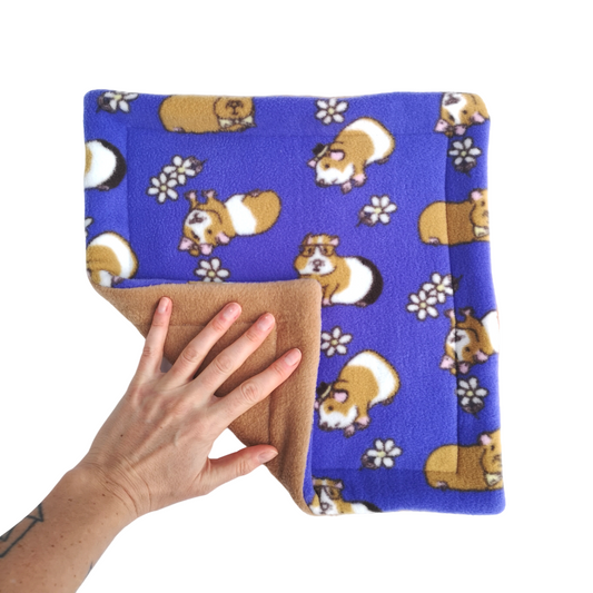 A hand holds a purple fleece pet pee pad featuring brown and white guinea pigs with a light brown reverse side