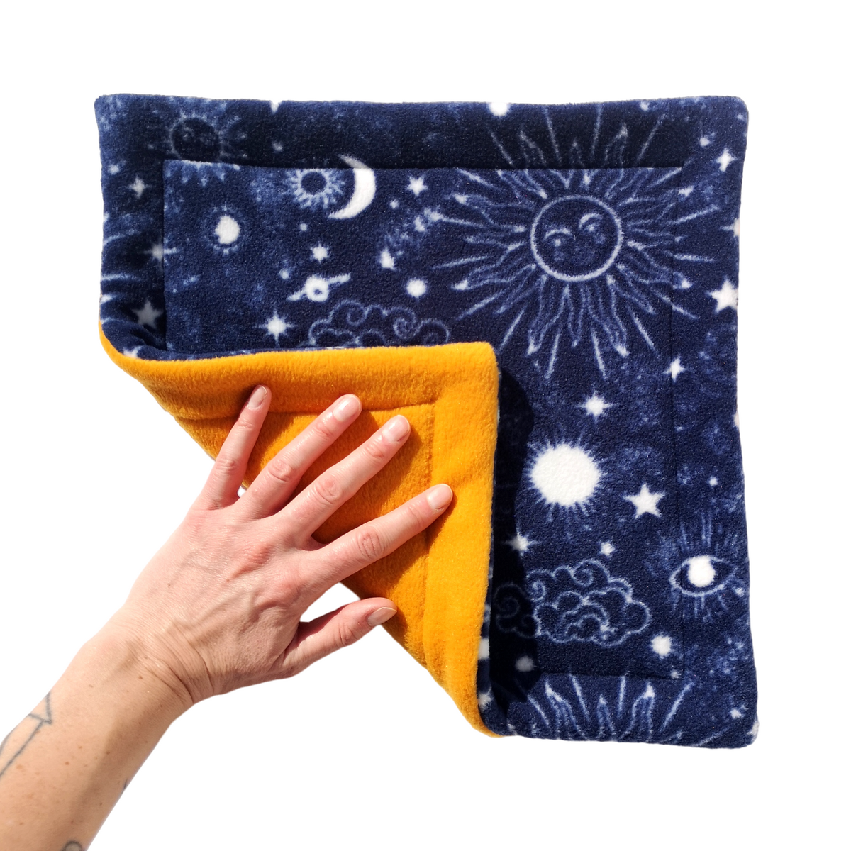 A hand holds a dark blue celestial themed print fleece pet pee pad featuring sun, moon and stars with a gold reverse side