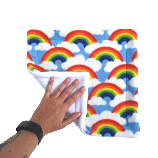 A hand holds a rainbows and clouds print fleece pet pee pad with a white reverse side